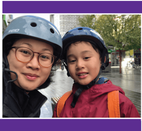 Child and adult wearing helmets for outdoor biking safety in the city, smiling at the camera.