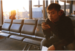 Young man using smartphone at airport terminal with city skyline view.
